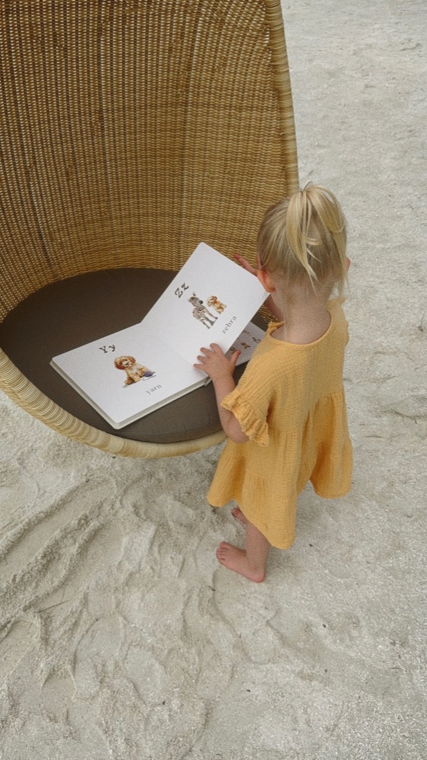 A small reader holding the ABC Doodle Dogs book in a wicker chair